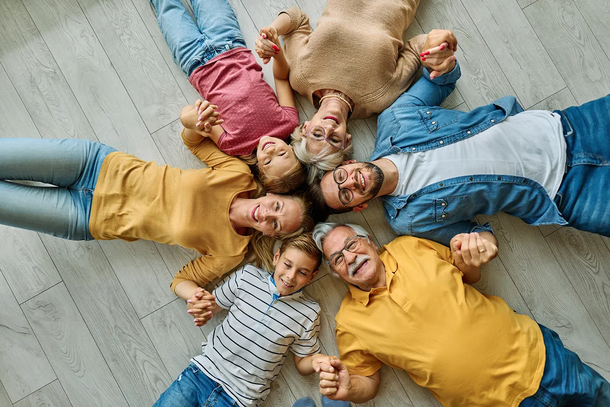 Drei Generationen einer Familie liegen lächelnd im Kreis auf dem Boden und halten sich an den Händen – Großeltern, Eltern und Kinder von oben fotografiert – Symbolbild für Familienpool