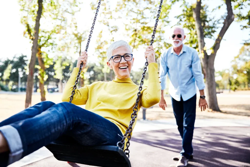 Älteres Ehepaar im Park – Frau lacht auf einer Schaukel, Mann schiebt sie an, Symbolbild für die Güterstandsschaukel