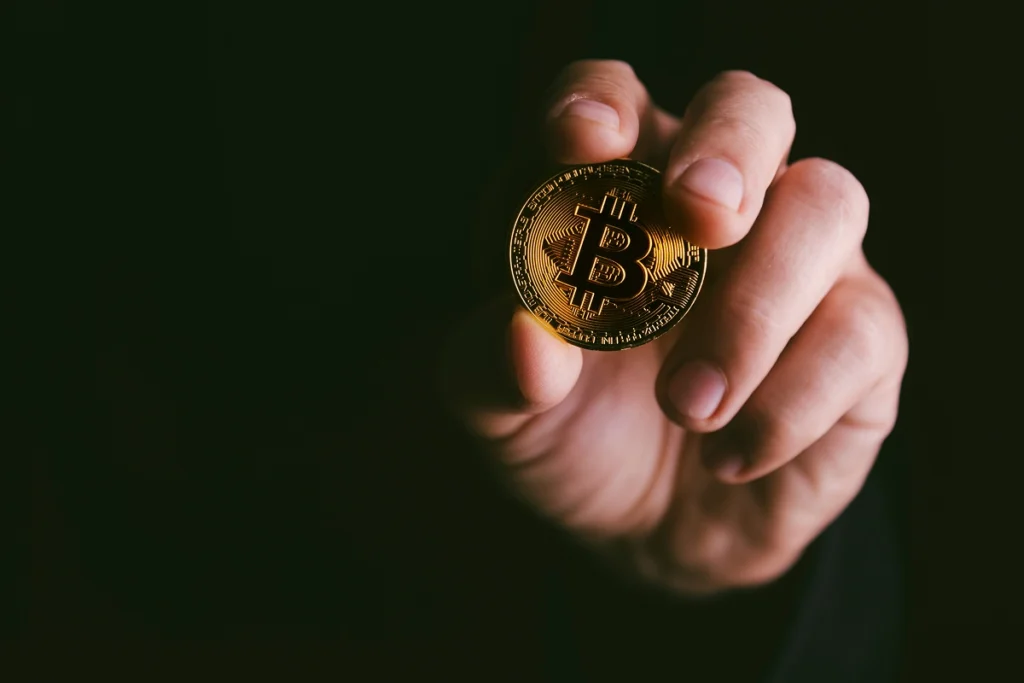 Hand hält eine goldene Bitcoin-Münze vor dunklem Hintergrund als Symbol für Kryptowährungen vererben und schenken.